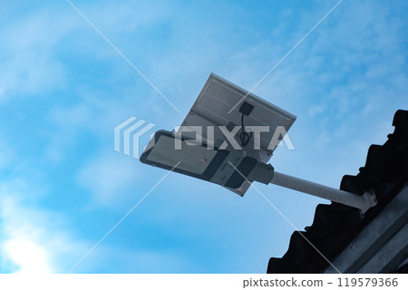 Mini solar cell panel and hd floodlight panal installed together under house eaves to store and use the power from the sunlight with street light and bulbs at night, sunset background, soft focus. 119579366