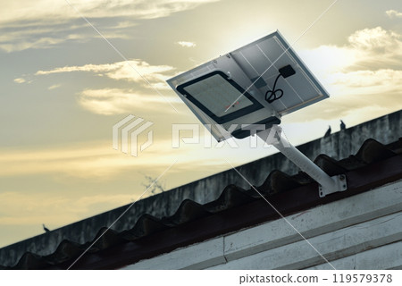 Mini solar cell panel and hd floodlight panal installed together under house eaves to store and use the power from the sunlight with street light and bulbs at night, sunset background, soft focus. 119579378