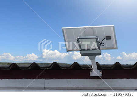 Mini solar cell panel and hd floodlight panal installed together under house eaves to store and use the power from the sunlight with street light and bulbs at night, sunset background, soft focus. 119579383