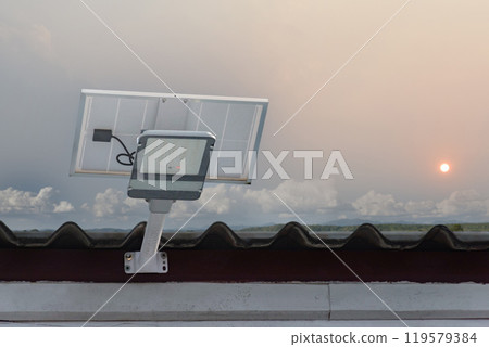 Mini solar cell panel and hd floodlight panal installed together under house eaves to store and use the power from the sunlight with street light and bulbs at night, sunset background, soft focus. Mini solar cell panel and hd floodlight panal installed together under house eaves to store and use the power from the sunlight with street light and bulbs at night, sunset background, soft focus. 119579384