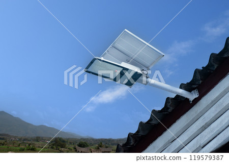 Mini solar cell panel and hd floodlight panal installed together under house eaves to store and use the power from the sunlight with street light and bulbs at night, sunset background, soft focus. Mini solar cell panel and hd floodlight panal installed together under house eaves to store and use the power from the sunlight with street light and bulbs at night, sunset background, soft focus. 119579387