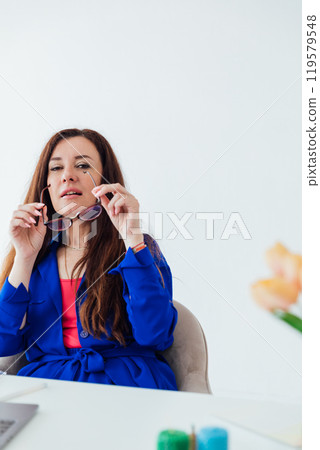 Brunette woman in blue business suit at desk in white office Brunette woman in blue business suit at desk in white office 119579548