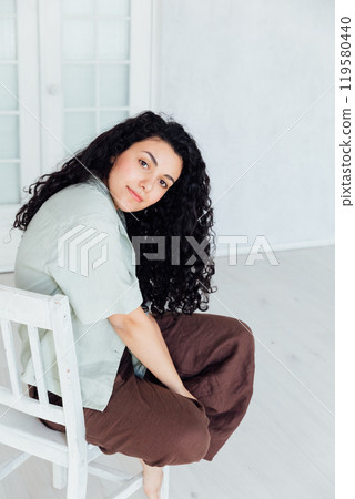 Curly brunette woman sitting on a chair at home Curly brunette woman sitting on a chair at home 119580440