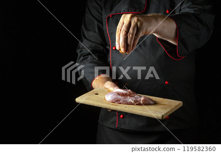 The cook sprinkles dry aromatic spices on a meat steak with his hand on a wooden board. The concept of cooking a meat dish by the hands of a chef on a black background 119582360