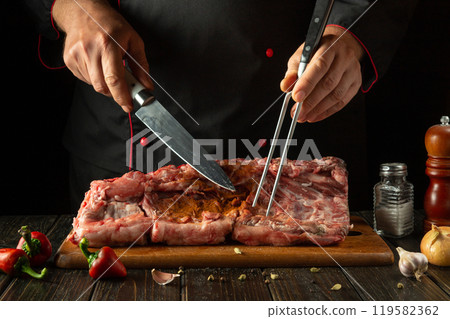 Chef preparing raw beef meat. Preparing a large piece of ribs before roasting. Fork and knife in the hands of a butcher on the kitchen table before cutting meat 119582362