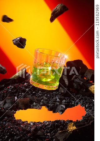 A glass of yellow cocktail floating suspended in the middle on a pebbles ring, with charcoal falling from above against a red-orange background, giving the impression of a powerful meteorite fall. A glass of yellow cocktail floating suspended in the middle on a pebbles ring, with charcoal falling from above against a red-orange background, giving the impression of a powerful meteorite fall. 119582969