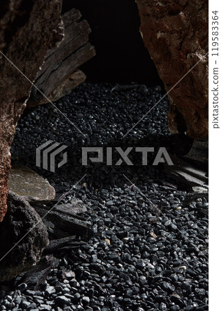 Pebbled surface with empty space in the middle, perfect for product display, surrounded by charcoal samples and large rugged rocks, set in a minimalist space. Ads image sample with high-angle view. 119583364