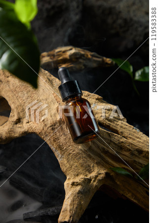 An amber glass dropper bottle set in the middle of a piece of weathered wood, surrounded by smoke and little pieces of charcoal and leaves, forms the photo-chic advertisement. 119583388