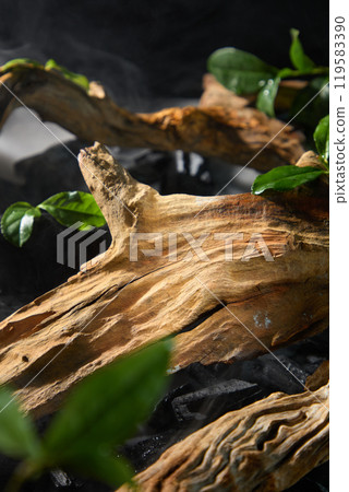 In the simple image, a piece of brown wood that is used to display the product is placed on a bed of black charcoal. There is smoke all around, and there are a few green leaves scattered. 119583390