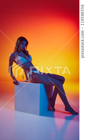 Woman, dressed white swimwear, leans back on white block, showing her slim figure in neon light against gradient studio background. 119586358