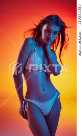 Photo of young woman in white bikini posing and her hair flowing on wind in neon light against gradient studio background. Photo of young woman in white bikini posing and her hair flowing on wind in neon light against gradient studio background. 119586389