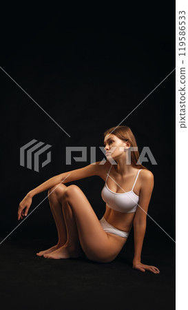 Slender young woman, dressed white lingerie posing, sitting , shows flexibility and balance against black studio background. 119586533