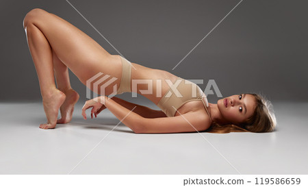 Young fit woman bridging her body upward, showing balance and strength, wearing neutral bikini against warm grey background. Young fit woman bridging her body upward, showing balance and strength, wearing neutral bikini against warm grey background. 119586659