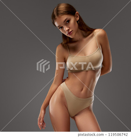 Pretty, beautiful woman posing, dressed beige lingerie, elegantly to emphasize curve of her slim, fit body against warm grey background. 119586742