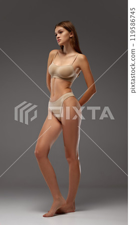 Young female model strikes thoughtful pose, showing off toned muscles against warm grey background. Full-body portrait. 119586745