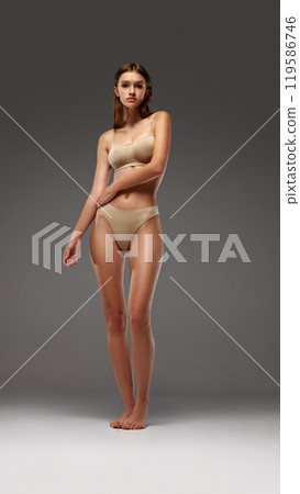 Slender, beautiful woman stands with slight lean, emphasizing her natural curves against warm grey background. 119586746