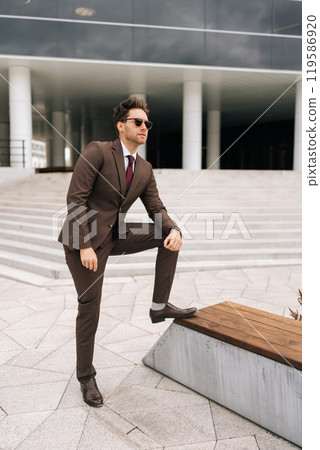 Vertical portrait of confident young businessman in stylish brown suit and sunglasses posing outdoors, exuding success and professionalism, city architecture complementing modern urban look. 119586920