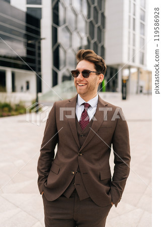 Vertical portrait of happy successful businessman wearing elegant suit and sunglasses standing posing outdoors in city with hands in pockets, smiling looking away. Modern cityscape backdrop background Vertical portrait of happy successful businessman wearing elegant suit and sunglasses standing posing outdoors in city with hands in pockets, smiling looking away. Modern cityscape backdrop background 119586928