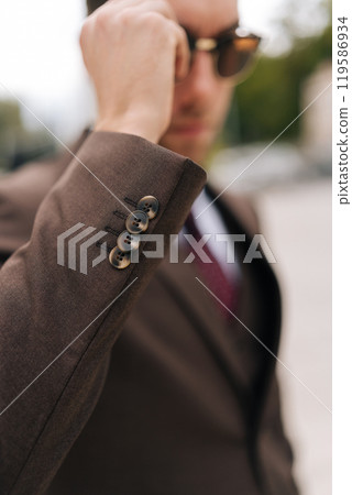 Selective focus shot of serious confident businessman adjusting sunglasses standing in front of modern office building, exuding professionalism and ambition, embodying success and leadership. Selective focus shot of serious confident businessman adjusting sunglasses standing in front of modern office building, exuding professionalism and ambition, embodying success and leadership. 119586934
