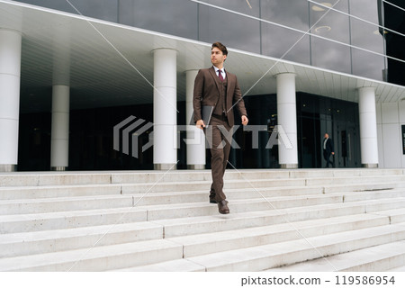 Portrait of confident businessman in suit descending office building stairs with laptop in hand, showcasing professional demeanor. Successful business man walking down stairs of modern office building 119586954