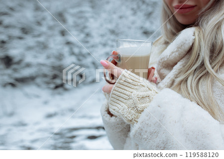 Beautiful hands hold a cup of cappuccino against the background of the winter forest. Gently beige gloves on the hands of a girl in the hands of coffee 119588120