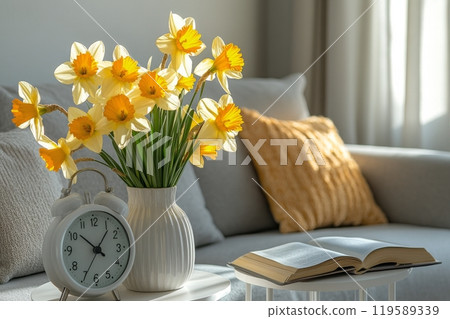 Bright yellow daffodils in a vase next to a cozy couch and an open book in a well lit living room 119589339