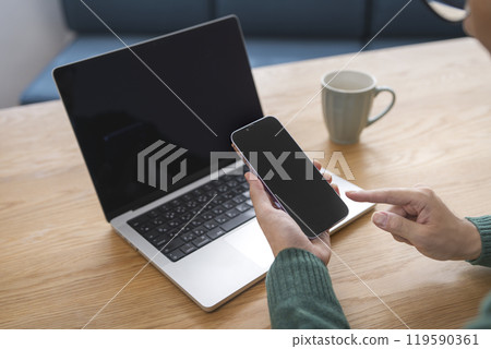 Hands of a man holding a smartphone in front of a laptop 119590361