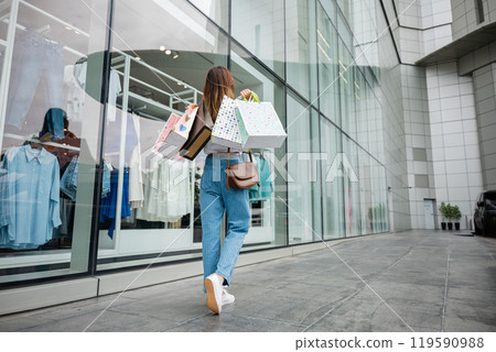 Back view of stylish woman holding shopping bags, walking in front of clothing store during the summer sale season. She is a fashionista, always on the lookout for the latest clothes and accessories 119590988