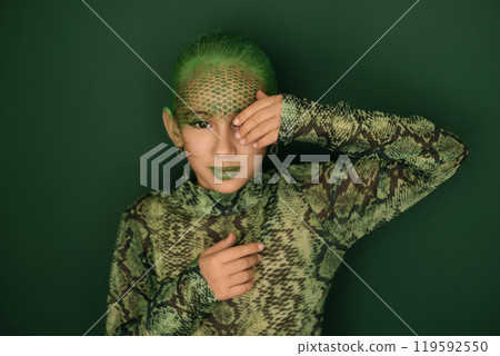 A sensual teenage girl in the guise of a snake, with a face painted with green paint, covers her eye with her hand. The symbol and mascot of the new year 2025. 119592550