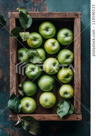 Fresh green apples arranged neatly in a rustic wooden crate with leaves on a dark background 119596533