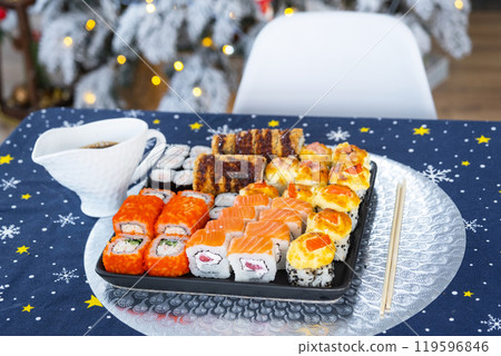 Delivery of sushi and rolls in food containers on the table in the festive interior of the kitchen of the house with a white Christmas tree for Christmas and New year. Food delivery for the holidays 119596846
