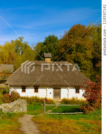 An old clay village house with a thatched roof and wooden shutters. Picturesque autumn landscape in the open-air ethnographic museum. Pirogovo Ukraine 119597142