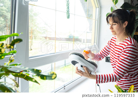 A woman launches a window cleaner robot of the house inside the interior with home plants. Household robotics, household assistant, cleaning servise 119597333