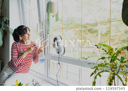 A woman launches a window cleaner robot of the house inside the interior with home plants. Household robotics, household assistant, cleaning servise A woman launches a window cleaner robot of the house inside the interior with home plants. Household robotics, household assistant, cleaning servise 119597343