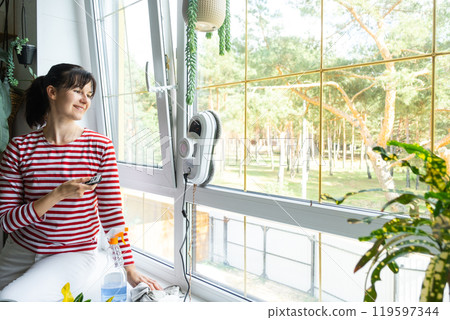A woman launches a window cleaner robot of the house inside the interior with home plants. Household robotics, household assistant, cleaning servise A woman launches a window cleaner robot of the house inside the interior with home plants. Household robotics, household assistant, cleaning servise 119597344