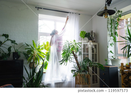 Woman hangs transparent tulle curtains on large windows in the house inside the interior. Spring cleaning, tidying up 119597573
