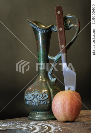 A knife is inserted into Naturally pink fresh kissabel apple fruit with Antique iron jug on wooden cutting board. A knife is inserted into Naturally pink fresh kissabel apple fruit with Antique iron jug on wooden cutting board. 119600469