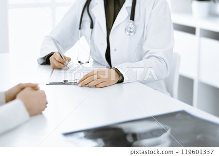 Unknown woman-doctor and female patient sitting and talking at medical examination in clinic, close-up. Therapist wearing green blouse is filling up medication history record. Medicine concept 119601337