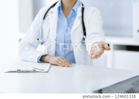 Unknown woman-doctor in blue blouse is giving helping hand while sitting at the desk in clinic. Perfect medical service, medicine concept Unknown woman-doctor in blue blouse is giving helping hand while sitting at the desk in clinic. Perfect medical service, medicine concept 119601338