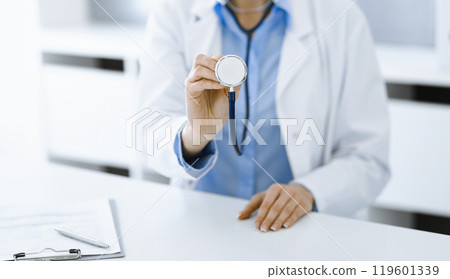 Unknown woman-doctor in blue blouse is holding stethoscope head while sitting at the desk in clinic, close-up. Physician ready to examine and help patient. Medicine concept Unknown woman-doctor in blue blouse is holding stethoscope head while sitting at the desk in clinic, close-up. Physician ready to examine and help patient. Medicine concept 119601339