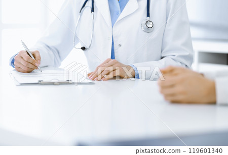 Unknown woman-doctor and female patient sitting and talking at medical examination in clinic, close-up. Therapist wearing blue blouse is filling up medication history record. Medicine concept Unknown woman-doctor and female patient sitting and talking at medical examination in clinic, close-up. Therapist wearing blue blouse is filling up medication history record. Medicine concept 119601340