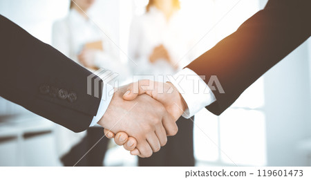 Businessman and woman shaking hands with colleagues at the background. Handshake at meeting in sunny office Businessman and woman shaking hands with colleagues at the background. Handshake at meeting in sunny office 119601473