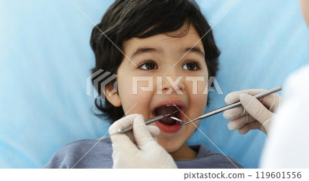 Cute arab boy sitting at dental chair with open mouth during oral checking up with doctor. Visiting dentist office. Stomatology concept Cute arab boy sitting at dental chair with open mouth during oral checking up with doctor. Visiting dentist office. Stomatology concept 119601556