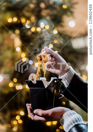 Hot noodles lifted with chopsticks from takeaway box under festive lights, creating cozy night vibe Hot noodles lifted with chopsticks from takeaway box under festive lights, creating cozy night vibe 119602488