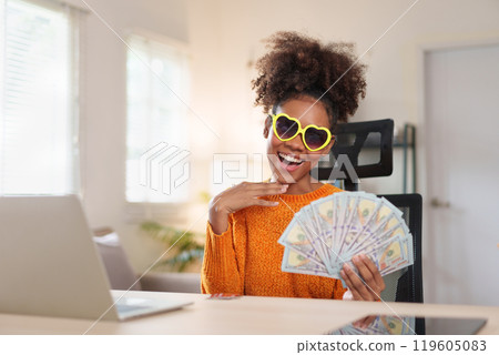 Joyful woman wearing heart-shaped sunglasses, holding a fan of dollar bills, smiling at the camera, expressing financial success and happiness. 119605083