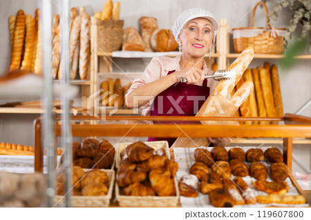 Elderly woman works as salesperson in bakery and packs baguettes in paper bag for customer 119607800