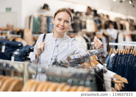 Middle-aged woman watching excitedly large stock of clothes in clothing store Middle-aged woman watching excitedly large stock of clothes in clothing store 119607833