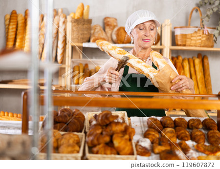 Elderly woman works as salesperson in bakery and packs baguettes in paper bag for customer 119607872