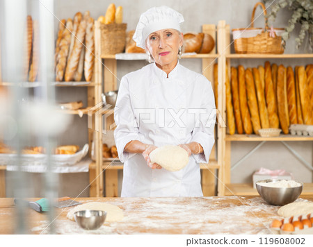 Elderly female chef skillfully kneads dough to bake delicious bread and croissants 119608002