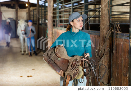 Woman jockey with saddle and bridle in her hands in stable Woman jockey with saddle and bridle in her hands in stable 119608003